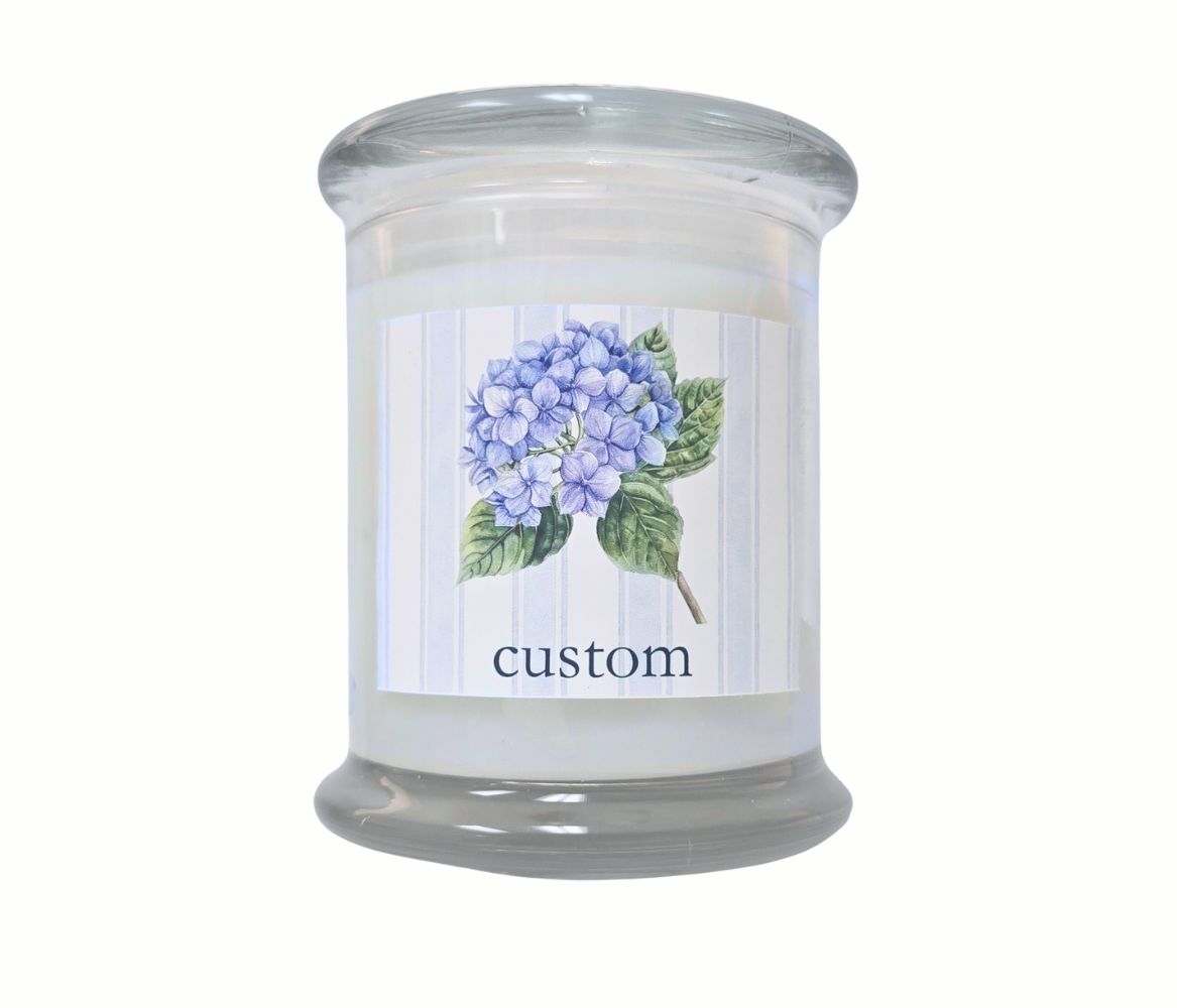 Personalized 12oz coastal candle with blooming hydrangea stem, soft blue striped background, and custom location label, hand-poured in eco-friendly coconut wax