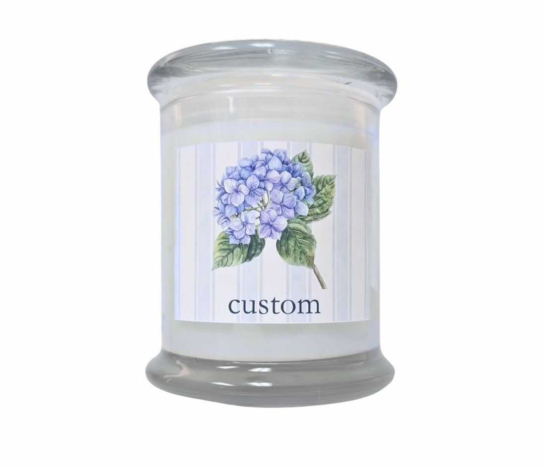Personalized 12oz coastal candle with blooming hydrangea stem, soft blue striped background, and custom location label, hand-poured in eco-friendly coconut wax