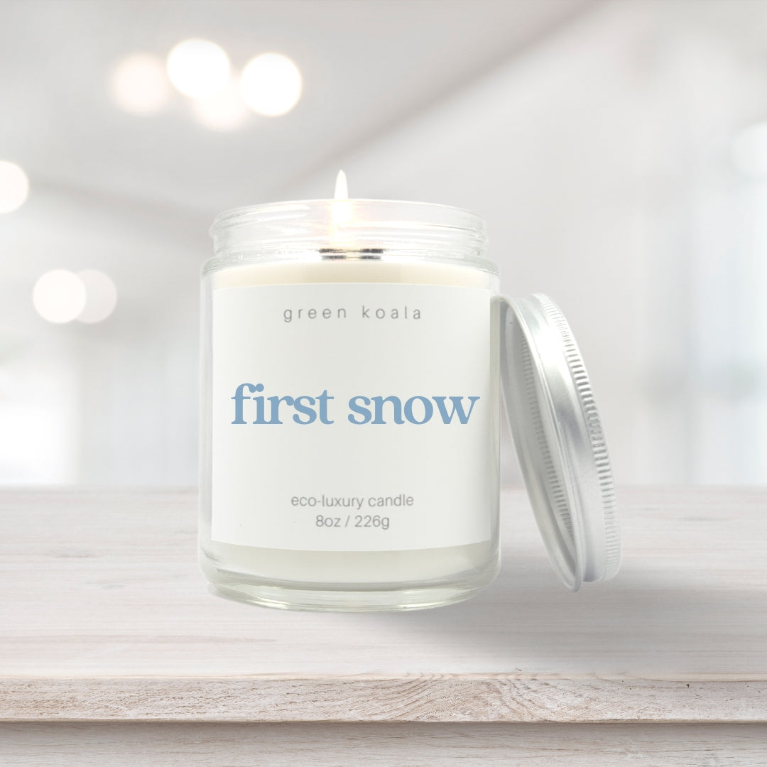 Image of First Snow candle in a glass jar with silver lid leaning on the side and burning on a wooden shelf. 