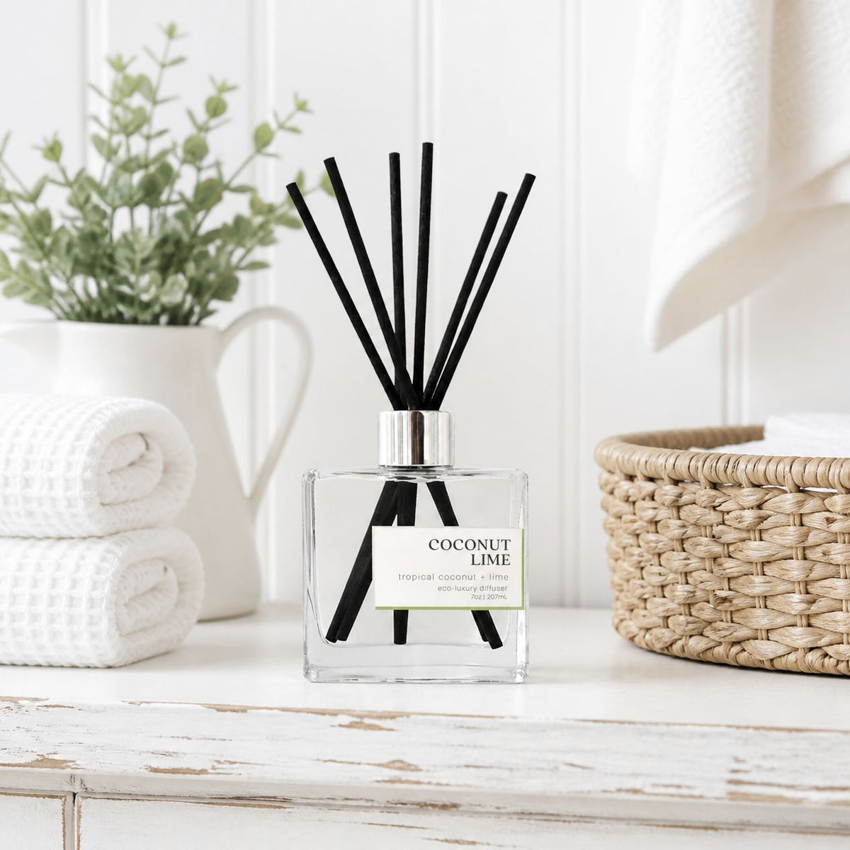 Coconut Lime non-toxic reed diffuser on a white rustic bathroom shelf with rolled towels, greenery and a woven basket