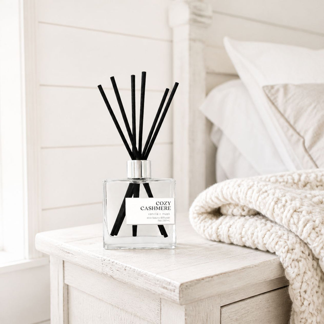 Cozy Cashmere non-toxic reed diffuser on a white farmhouse nightstand beside a cream chunky-knit throw