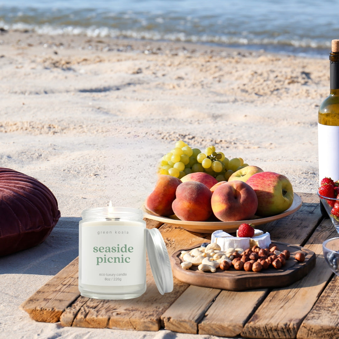 Image of Seaside Picnic candle in a glass jar with silver lid burning on a table with a fruit plate and wine on the beach. 