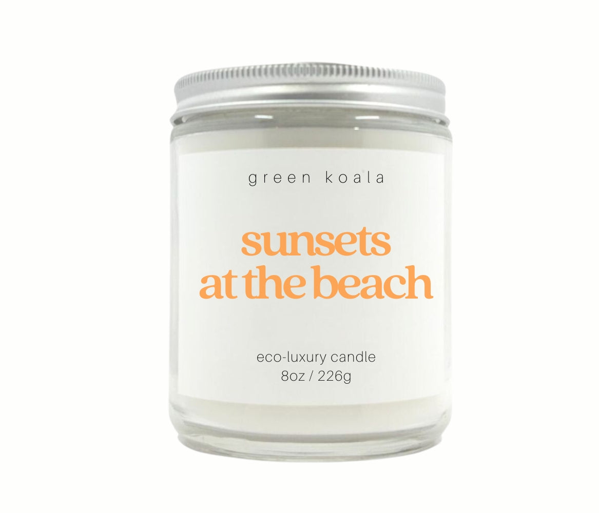 Image of Sunsets at the Beach candle in a glass jar with silver lid with a white background.  The text on the label is orange color. 